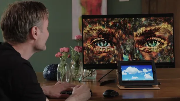 Een foto van Jeroen van der Most die links zit achter een tafel. Op het beeldscherm rechts op de tafel is een door de computer gemaakt kunstwerk te zien van twee groene ogen met wenkbrauwen en gekleurde vakjes eromheen in zwart, bruin, groen en rood.