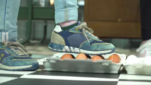 Een foto van een broek en schoenen. De rechterschoen staat op de bovenkant van acht bruine eieren in een open doos, de andere schoen nog op de grond. Het is een test hoe sterk de eieren zijn. De vloerbedekking heeft zwarte en witte vierkanten.