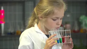 Een foto van Indi die een doorzichtig rekje vastheeft. Ze blaast in één van vijf glazen buisjes met vocht. Het vocht is geel, blauw, groen en rood en er zit in ieder buisje steeds iets meer vocht. De vijfde kleur is niet te zien door haar vingers. 