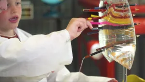 Een foto van Siem en linksboven de neus van Evelien, die een witte jas dragen. Evelien's arm steekt vanaf links een roze potlood door een doorzichtige plastic zak met water. Er zitten al acht andere potloden in de zak en uit één punt lekt er water uit.