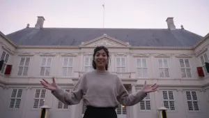 Een foto van presentator Manon die met haar armen wijd staat voor Paleis Noordeinde. Paleis Noordeinde is het het werkpaleis van Koning Willem-Alexander en Koningin Máxima.