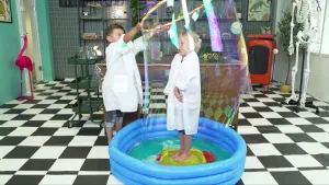 Een foto van Tijn en Sophie in een witte jas. Sophie staat in een blauw, opblaasbaar zwembadje. Tijn heeft een grote hoepel vast waar een enorme bel zeepsop aan de onderkant hangt.  De zeepsopbel zit helemaal om Sophie heen, die er lachend doorheen kijkt.