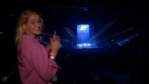 Een foto van presentator Janouk die in de camera kijkt met opgestoken duimen bij een groot optreden. Op de achtergrond is er publiek te zien die in het donker staan en is er een verlicht podium te zien.