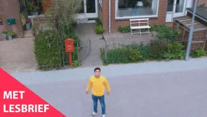 Een foto van de presentator in een straat. Je kijkt van boven op hem neer. Hij kijkt in de camera. Achter hem zie je een tuintje met een rode brievenbus. Linksonder in beeld staat de tekst "Met lesbrief".