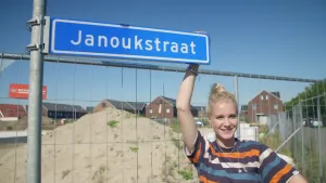 Een foto van Janouk die lachend naar de camera kijkt. Ze heeft met haar rechterhand een blauw straatnaambord vast boven haar hoofd met de straatnaam "Janoukstraat" erop. Het bord zit aan een stalen paal vast. Erachter staan hekken voor een berg zand.