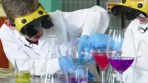 Een foto van Calvin en Sybren in een wit pak en gele bril met opengeklapte zwarte klepjes. In een maatbeker met paarse vloeistof giet er één lichtblauwe vloeistof. Ook een maatbeker met gele vloeistof en glazen met rode en paarse vloeistof zijn te zien.