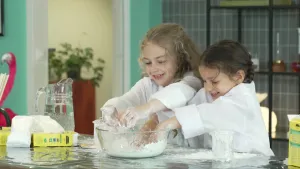 Een foto van Selina en Alyssa in een witte jas die een soort kneedbaar slijm maken: oobleck. Voor ze op tafel staat een glazen kom met wit spul erin: water uit een waterkan en maïzena uit een pak. Alyssa is na Selina bezig te kneden in de schaal. 