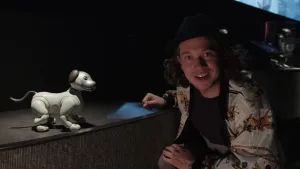 Een foto van Pascal die gehurkt naast een donker podium zit. Op het podium staat een grijze robothond, die van de zijkant te zien is. Hij heeft een blauw oog, bruin oor, bruine staart en zwarte neus. Pascal draagt een blouse met paarden en ruiters erop. 