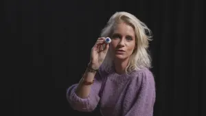 Een foto van presentator Janouk die in de camera kijkt en een kunstoog laat zien die zij in haar hand heeft.