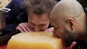 Een foto van Tim en Edson die likken aan een heel groot rechthoekig oranje blok op een rode tafel. Tim staat achter de tafel, Edson's gezicht is rechts in beeld. Het blok is een supergrote winegum, zoals het snoepje, en is door Tim en Edson gemaakt.