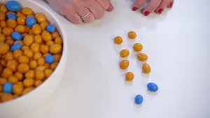 Een foto van een witte tafel. Op de tafel is links een witte schaal met blauwe en oranje M&M chocoladesnoepjes te zien. Aan de rechterkant liggen tien losse M&M snoepjes in rijen van twee: acht oranje en twee blauwe snoepjes. 