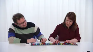 Een foto van Diederik en Ionica aan een witte tafel. Voor hen ligt een wit apparaatje waar een piano aanzit die bijna zo plat is als een vel papier. De piano heeft zwarte toetsen met toetsen van zeven verschillende kleuren ertussen. Ze spelen op de piano.