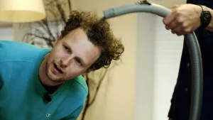Een foto van Tim. Hij heeft een lichtblauwe trui aan en staat gebukt. Iemand waarvan de blouse en hand zichtbaar zijn, houdt een grijze slang van een stofzuiger op zijn gekrulde, donkerblonde haar. Een pluk haar wordt opgezogen in de stofzuigerslang.