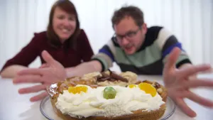 Een foto van stukken taart in een cirkel op een glazen bord. Een stuk slagroomtaart, een stuk appeltaart, een stuk mokkataart en een stuk notentaart zijn te zien. Eén van de twee personen achter de tafel houdt zijn beide handen naast het bord.