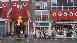 Een animatie van de foto van een normale winkelstraat met acht rode, stoffen doeken op de muren met hakenkruizen van de nazi's erop. Voor een winkel, de "Adolf Fan Store", hangen rode nazi vlaggetjes. Met witte lijnen getekende mensen dragen nazi vlaggen.