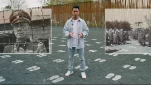 Een foto van een presentator in kamp Amersfoort. Op de grijze grond staan witte voetstappen voor ieder slachtoffer van de Holocaust op deze plek. Er staat een schutting van palen en prikkeldraad en links en rechts foto's van nazi soldaten in het kamp.