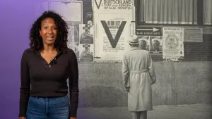 Een foto van een presentator die links staat en een zwart-wit foto rechts. Op de zwart-wit foto staat een man voor een muur met aanplakbiljetten. Te lezen is "Duitschland wint voor Europa op alle fronten" en een pijl naar rechts met "naar schuilplaats".