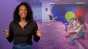 Een foto van een presentator die naast een foto van een ballenbak staat die zo groot is als een binnenzwembad, met een blauwe glijbaan ernaast en vol met roze ballen. Twee meisjes zitten aan de kant met hun voeten erin, één jongen springt erin.