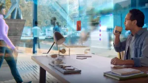 Een foto van een moderne wereld. Je ziet een man aan een bureau zitten. Hij drinkt koffie. Voor hem hangen doorzichtige beeldschermen in de lucht. Daarop zie je teksten en plaatjes. Op de achtergrond loopt een vrouw voorbij. 