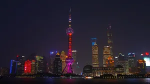 Een foto van het uitzicht op een grote stad. Op de voorgrond zie je een rivier. Daarachter hele hoge gebouwen. Het is donker, maar die gebouwen zijn verlicht in verschillende kleuren.