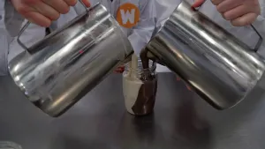 Een foto van twee mensen in witte jas met "W" erop achter een tafel, hun gezicht is niet te zien. Ze zijn allebei chocopasta aan het schenken in één glazen potje uit een schenkkan van staal. Links loopt de witte pasta in het potje, rechts de bruine pasta.