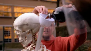 Een foto van Tim met een plastic masker achter een voorbeeld van een menselijk skelet, waarvan het hoofd en de nek te zien zijn. Naast de nek steekt een plastic buis door het skelet. Tim giet er cola uit een fles in met een trechter, een doorgietbuisje.