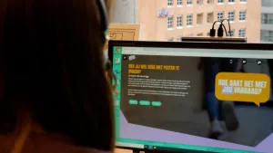 Een foto van iemand achter een beeldscherm. Op het scherm is "Heb jij wel eens met pesten te maken?" te lezen en een tekstballon met "hoe gaat het met jou vandaag?". De website is niet te lezen, maar het is een pagina op kindertelefoon.nl.