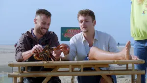 Een foto van presentator Jurre die buiten met zijn been op tafel zit en naar een kreeft kijkt die de man die naast hem zit vasthoudt. 