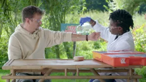 Een foto van een vrouw die water uit een kan giet over de arm van presentator Jurre.
