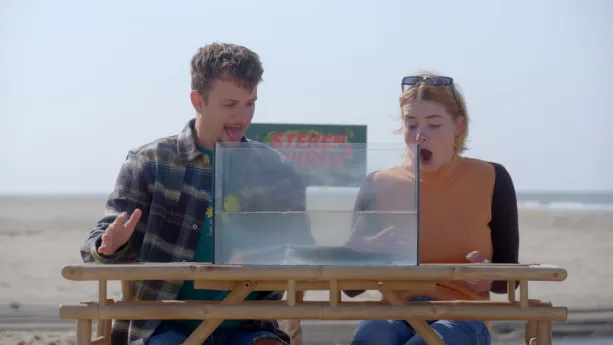 Een foto van presentatoren Jurre en Emma die met open monden naar een bak met water kijken waar een paling in zit. Een paling is een vis die op een slang lijkt.