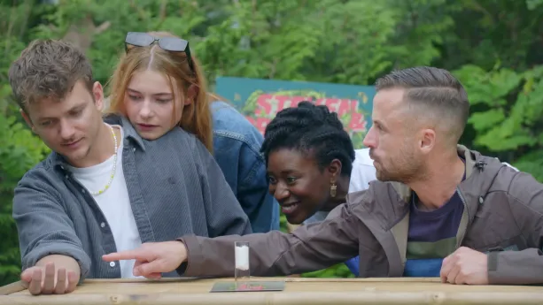 Een foto van presentator Jurre die naar iets op zijn rechterarm kijkt. Presentator Emma kijkt mee terwijl ze met haar kin op zijn schouder leunt. Er wijst een man naar de rechterarm van Jurre en er kijkt nog iemand mee die tussen hun in staat.