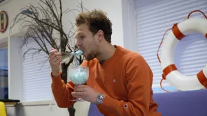 Een foto van Tim die een ijsje uit een groot glas eet. Het ijsje heeft een lichtblauwe kleur en is een door Tim en Edson zelfgemaakt smurfenijsje. Tim neemt een hap van een lepel. Tim zit op een paarse bank met op de rugleuning een witte reddingsboei.