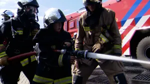Een foto van drie mannen en een jongen, Sam, in een brandweerpak met gele en zilveren strepen erop en met een brandweerhelm op. Sam heeft een rode brandweerslang vast, waar water uit spuit naar rechts. Op de achtergrond staat een rode brandweerauto.