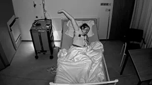 Een zwart-wit foto van Louisa die in een ziekenhuisbed ligt op haar linkerzij met de linkerarm in de lucht. Ze heeft meetapparatuur op haar borst hangen en er lopen slangetjes naar haar neus en draadjes naar haar voorhoofd voor een slaaponderzoek.