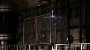 Een foto van Tim en Taha die in een stalen kooi van Faraday, die beschermt tegen elektriciteitsschokken, zitten op een stoel. Boven de kooi komt een kronkelende bliksemschicht van vijfhonderdduizend volt, wit met een paarse rand, door een buis heen. 