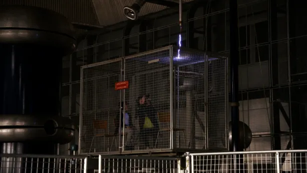 Een foto van Tim en Taha die in een stalen kooi van Faraday, die beschermt tegen elektriciteitsschokken, zitten op een stoel. Boven de kooi komt een kronkelende bliksemschicht van vijfhonderdduizend volt, wit met een paarse rand, door een buis heen.