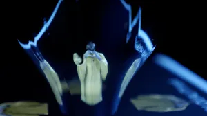 Een foto van Edson in een gele trui en met zwarte pet op die langwerpig en een beetje misvormd naast twee kopieën van hem staat die schuin te zien zijn op een laptopscherm. De camera laat tegelijk Edson en een drinkglas op het scherm van de laptop zien.