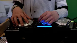 Een foto van een mengpaneel met twee grote, zilverkleurige draaiknoppen, stekkerpunten voor kabels, knopjes en een blauw oplichtend, rechthoekig scherm. Achter de draaitafel zijn de handen van de DJ te zien, die aan de knop links in beeld draait.