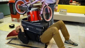 Een foto van Edson die op een plank met lange spijkers, zoals van een spijkerbed, op de grond ligt. Zijn hoofd ligt op een rood kussen. Op zijn buik liggen voorwerpen opgestapeld: een blauwe koffer, een rode trommel, een roze kinderfiets en drie boeken.