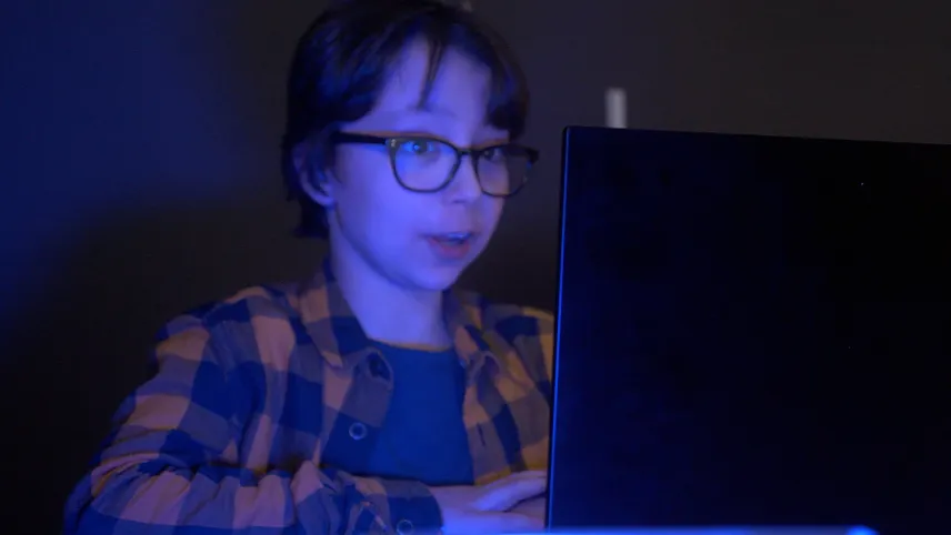 Een foto van de jongen Storm, die achter een groot, zwart beeldscherm zit. Hij wordt verlicht door het blauwe licht van het beeldscherm. Hij draagt een geel met zwart geruite blouse en een donker t-shirt eronder. Ook heeft hij een donkerkleurige bril op.