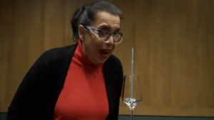 Een foto van een vrouw die aan het zingen is met haar mond open. Ze draagt een veiligheidsbril voor haar gewone, zwarte bril. De vrouw zingt in de richting van een wijnglas. In het glas staat middenin een blauw, plastic rietje, dat recht omhoogsteekt.