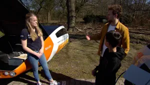 Een foto van een vrouw voor een auto die werkt op zonne-energie bij de Technische Universiteit Eindhoven. De auto is oranje en wit van kleur met een zwart, bol dak. De motorkap staat open en er ligt een witte accu in. Elias en de presentator staan rechts.
