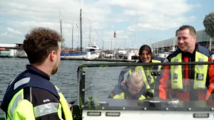 Een foto van de voorruit van een boot in het water en achteraan boten die stilliggen. Achter de ruit staan twee volwassenen en een kind die een lichtgeel opblaasbaar vest met zilverkleurige strepen dragen en links voor de ruit een man met hetzelfde vest.