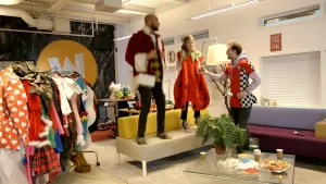 Een foto van een ruimte met rechts twee banken en een glazen tafel, links een rek met kleurige kleding en achteraan een bureau. Een man in een kerstmanjas en een meisje in een aardbeikostuum springen op de linkerbank, de presentator springt vóór de bank.