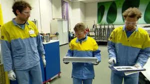 Een foto van Tim, Thijs en Matthijs, de medewerker van een staalfabriek, die een blauwe broek, geel met blauwe blouse en een veiligheidsbril dragen. Thijs heeft een rechthoekig stuk staal vast, Tim een klembord met bovenop een papier vol foto's en tekst.