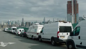 Een foto van een rij geparkeerde auto's, bestelbusjes en een kampeerauto. Op de achtergrond zie je het uitzicht op een grote stad, met veel hoge gebouwen. 