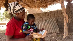 Een foto van een Afrikaanse vrouw met een hoofddoek. Ze zit buiten, op de grond, onder een rieten dak. Ze heeft haar kind op schoot en houdt een boek vast. Het kind kijkt aandachtig naar dat boek. 