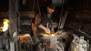 Een foto van een smid in een smederij. De smid heeft een baard, en een muts op. Hij is aan het werk. Hij slaat met een hamer op iets heets. De vonken spatten eraf. Om hem heen zie je veel gereedschap.