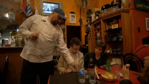 Een foto van een Russisch eetcafé in Argentinië. Je ziet een tafel met twee mensen eraan. Naast de tafel staat een bediende met een wit hemd aan. Hij wijst naar een smartphone die op hun tafel ligt. Alle drie kijken ze naar dat schermpje.
