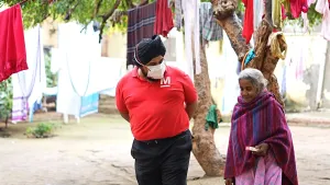 Een foto van een man en vrouw in India. De man draagt een tulband en een mondmasker. Ze lopen op straat en zijn met elkaar in gesprek. Op de achtergrond zie je waslijnen met kleren eraan. 